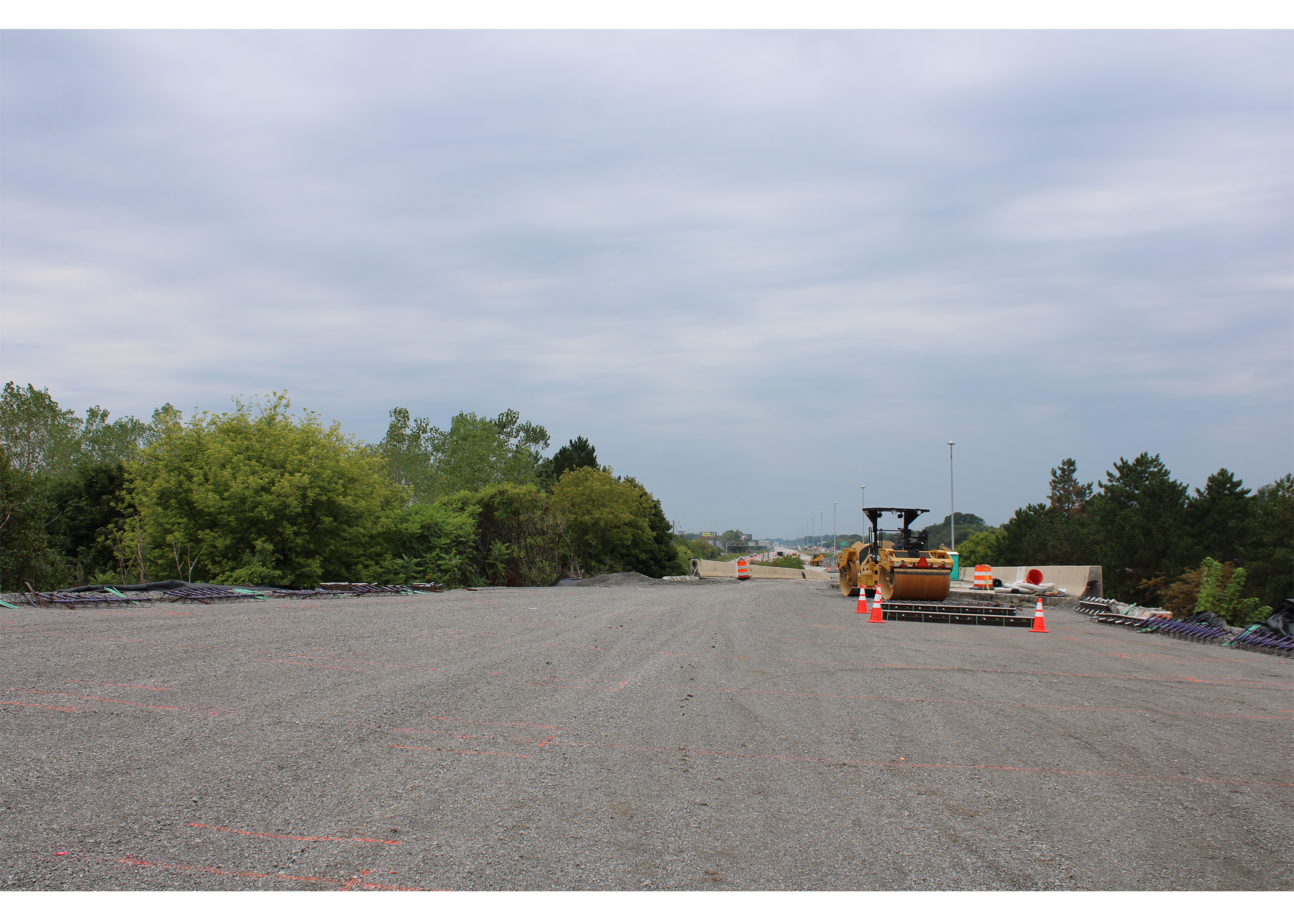 Wide stretch of new gravel roadbed marked with orange survey paint. A road roller and traffic cones sit on the right, with trees lining both sides of the corridor.
