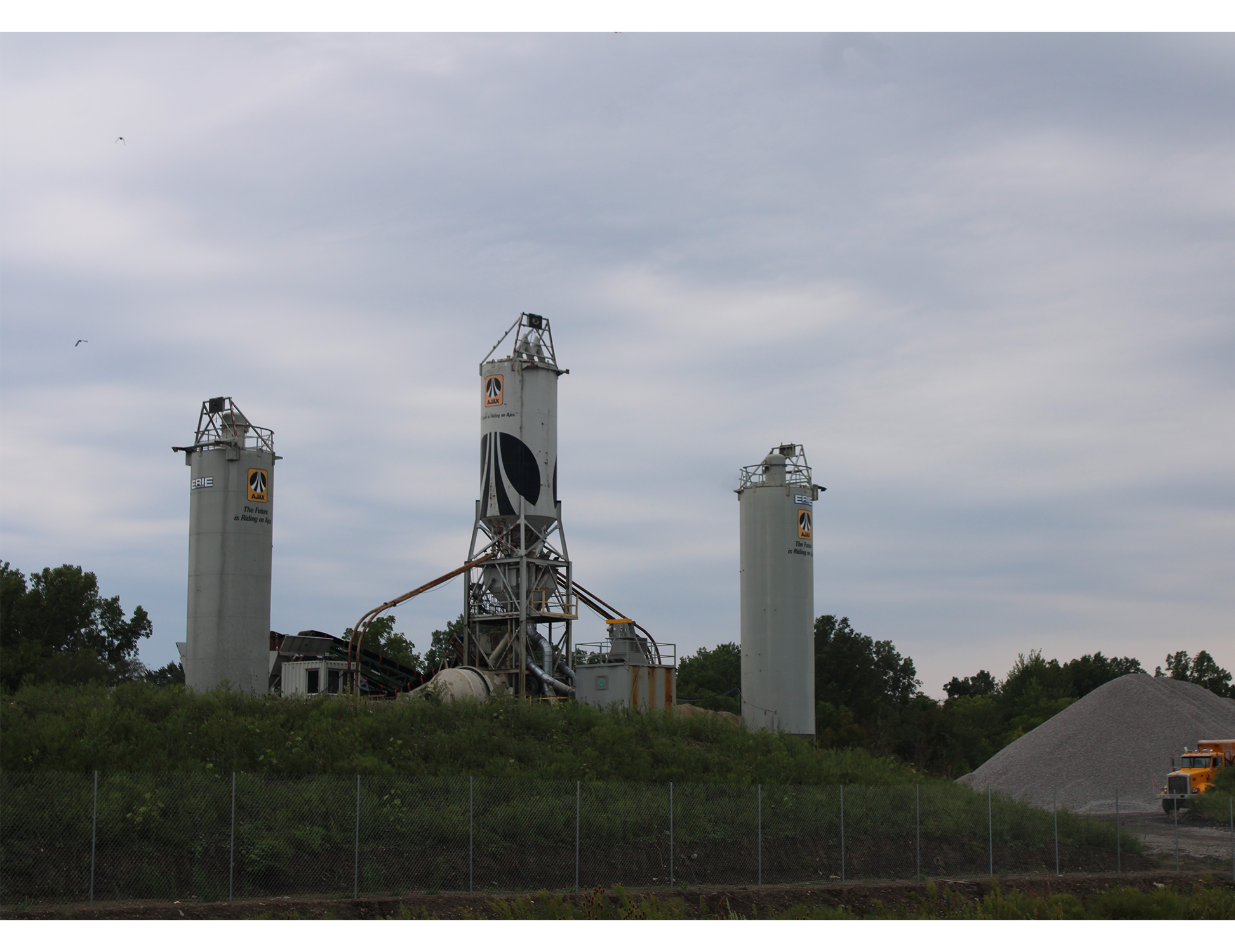 Concrete batch plant with three tall silos standing behind a chain-link fence. A large gravel pile and an orange dump truck sit at the right.