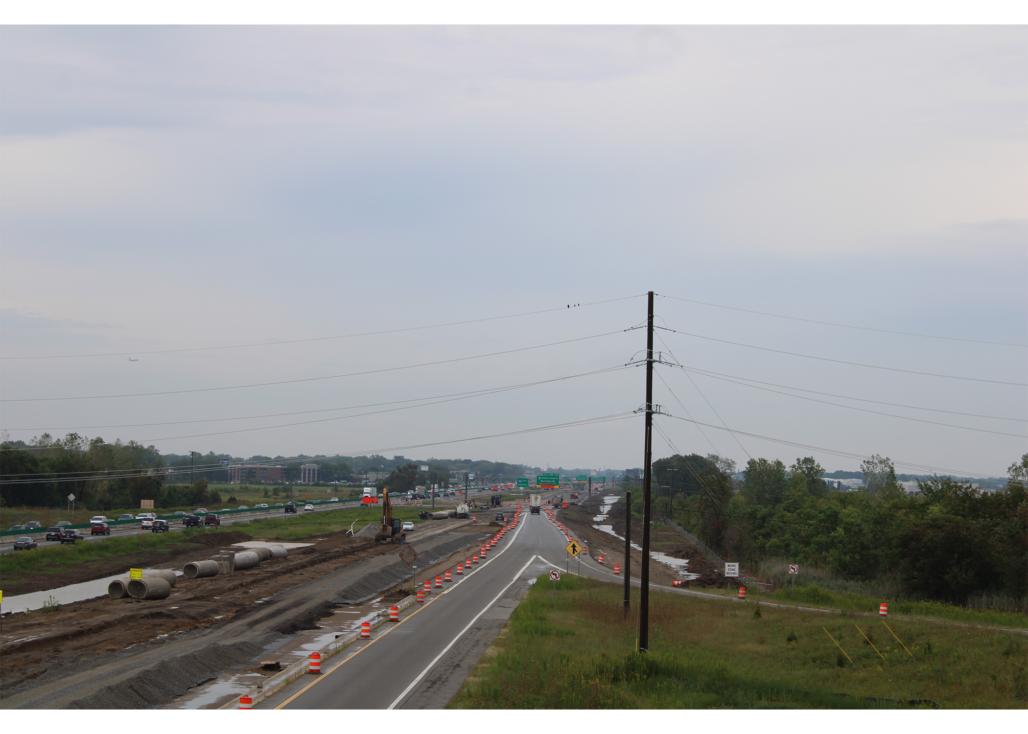 High view down the I-94 corridor. Traffic is shifted into a single barrel-lined lane beside an excavated area with drainage pipes and an excavator, with overhead exit signs visible far ahead