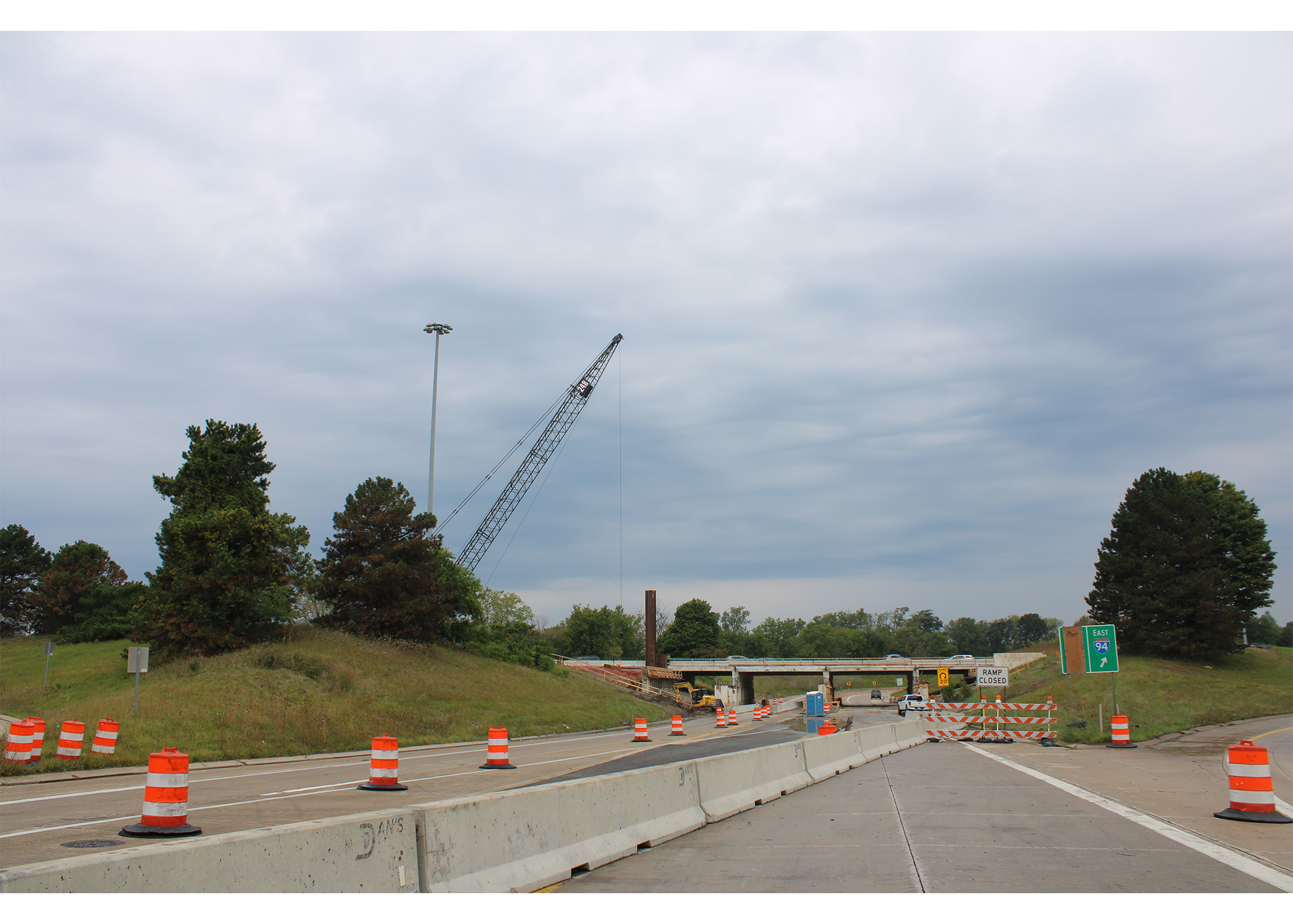 Closed on-ramp to eastbound I-94 blocked by striped barricades and a "Ramp Closed" sign. Concrete barriers and orange barrels line the road, and a crawler crane rises beside an overpass under construction.