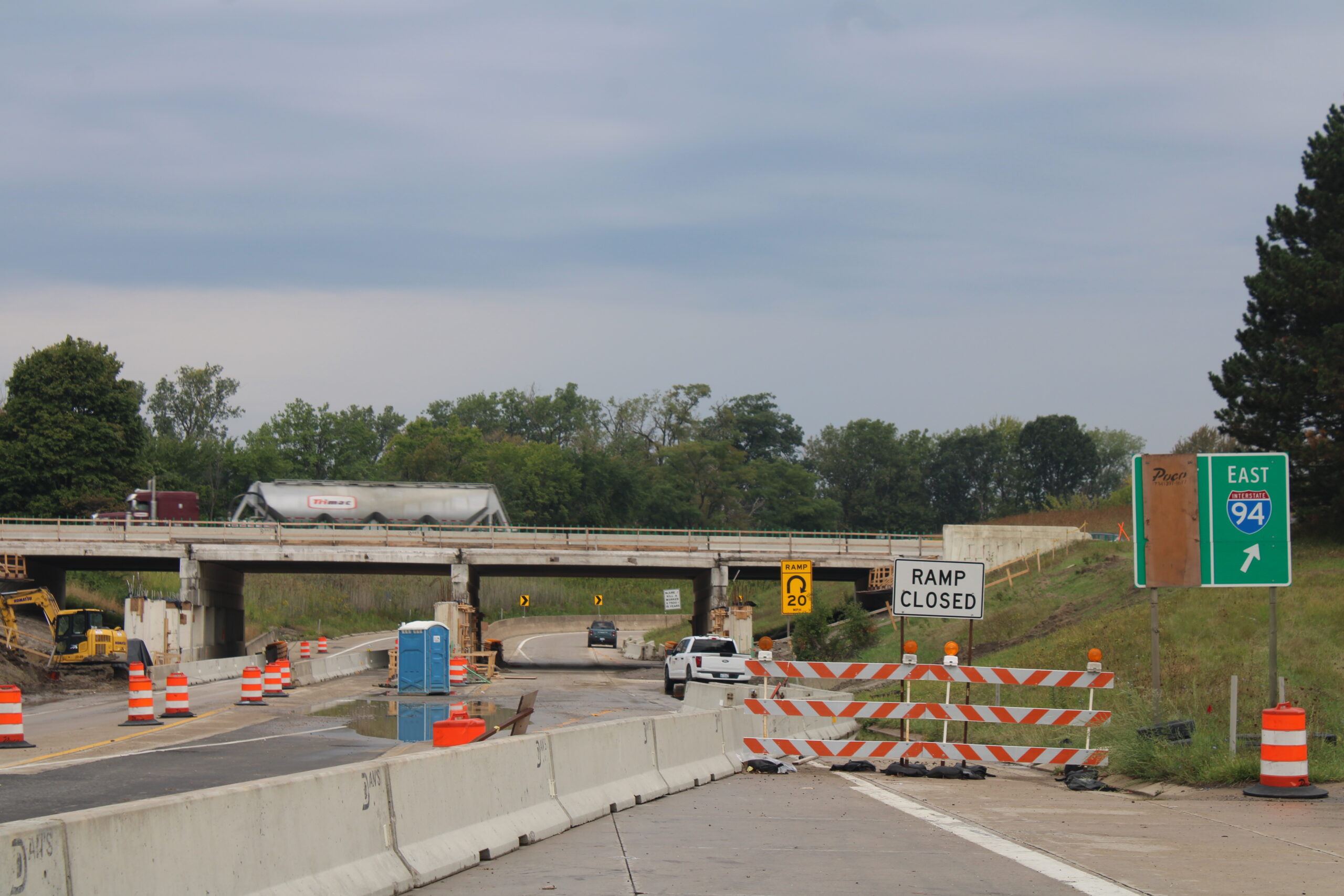 Close view of a closed ramp to I-94 East with barricades, a "Ramp Closed" sign, and a 20 mph ramp advisory sign. Beyond it, a tanker truck crosses an overpass whose piers are being rebuilt, with a portable toilet and excavator nearby.