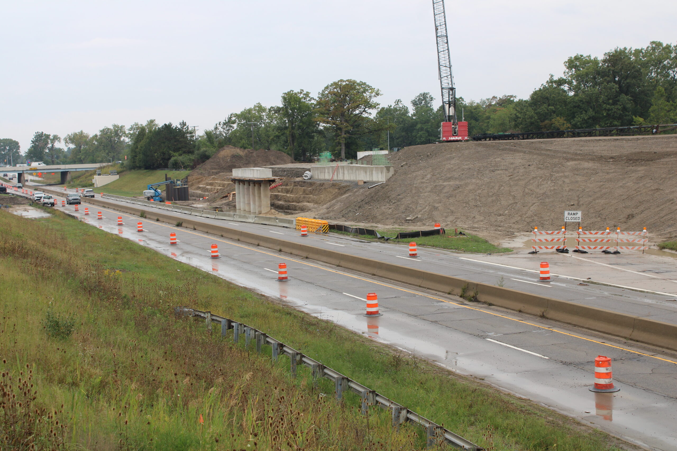 Elevated view of wet, barrel-lined freeway lanes beside a graded embankment. A new bridge pier and abutment wall are under construction, a crane sits atop the hill, and barricades block a closed ramp.